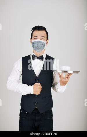 Handsome happy young waiter in medical mask carrying cup of coffee on metal tray Stock Photo