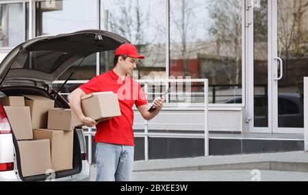 Online ordering and transportation. Courier looks at tablet and holds box near entrance Stock Photo