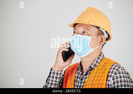 Asian construction worker in hardhat and medical mask calling on phone Stock Photo