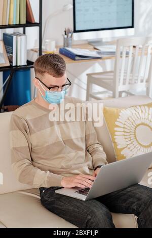 Software developer in medical mask sitting on sofa at home at working on laptop during lockdown Stock Photo