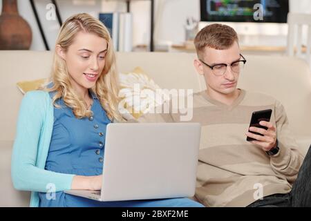 Smiling young woman sitting on sofa at home, answering e-mails from colleagues and clients when husband checking social media on smartphone Stock Photo