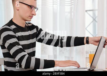 Young man in glasses sitting at kitchen table with glass of orange juice and opening laptop when starting new day of remote work Stock Photo