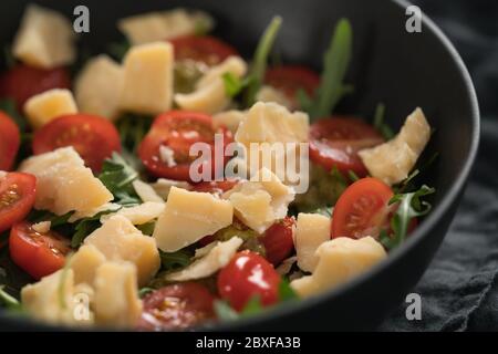 Fresh salad with cherry tomatoes, arugula and hard vintage cheese in black bowl on linen napkin Stock Photo