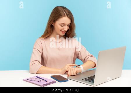 Positive smiling woman employee looking at watch on her hand, working on laptop in office, checking hour to take break, optimistic having enough time Stock Photo