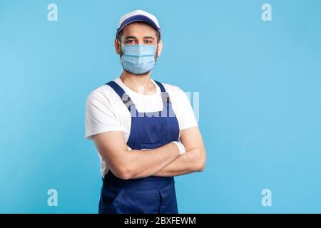 Portrait of happy handyman in overalls mask, and cap standing with crossed hands, profession of service industry, courier delivery. Expert house repai Stock Photo