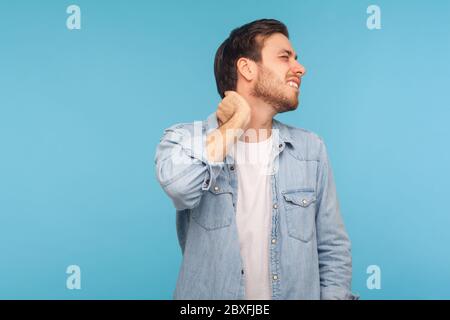 Portrait of exhausted young man in denim shirt touching neck and grimacing from intense pain, massaging to relieve muscle tension, hurting shoulders. Stock Photo