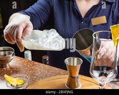 The barman in gloves holds in his hand a scoop with large pieces of ice and a stainless steel shaker. Workplace of the bartender. Making a soft drink Stock Photo