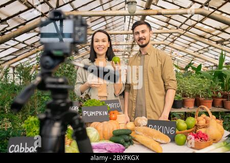 Positive young multi-ethnic couple showing apple to camera while shooting video review in greenhouse Stock Photo