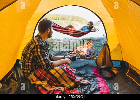 couple resting near fire. man sitting in tent woman laying on hammock Stock Photo