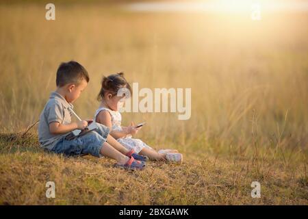 Two children sitting in a field playing on a tablet and mobile phone at sunset, Thailand Stock Photo