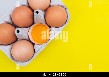A package of six brown eggs with one opened and orange yolk on yellow background with copyspace Stock Photo
