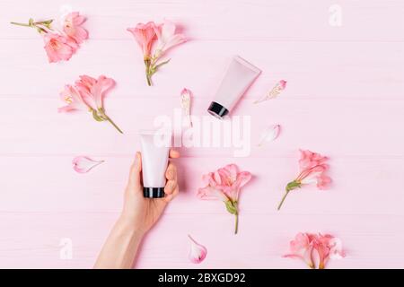 Woman's hand holding  tube of cosmetic cream among delicate flowers on pink background, flat lay. Stock Photo