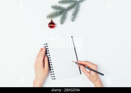 Woman's hands write in an empty notebook next to spruce branch with red Christmas tree ball on white background. Stock Photo