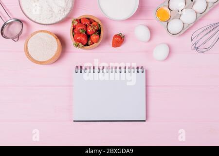 Ingredients for baking strawberry pie next to an empty notebook on pink table, flat lay composition. Stock Photo