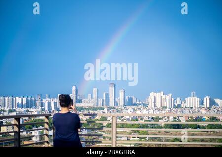 Young indian girl wearing hair in a bun and with blue dress photographing rainbow over gurgaon delhi noida cityscape on a monsoon day Stock Photo