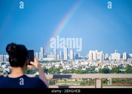 Young indian girl wearing hair in a bun and with blue dress photographing rainbow over gurgaon delhi noida cityscape on a monsoon day Stock Photo