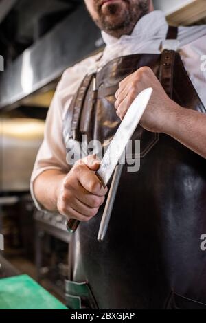 Close-up of butcher in leather apron sharpening blade of kitchen knife using honing rod Stock Photo