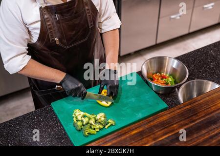Above view of unrecognizable chef in leather apron standing at counter and cutting bell pepper on plastic board Stock Photo