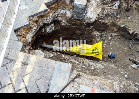 A hole in the pavement with an electrical conduit, a yellow plastic wrapper over the end reading 'Danger: Live Cable', London, UK Stock Photo