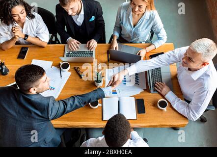Businessmen Handshaking During Corporate Meeting Sitting At Table In Office Stock Photo