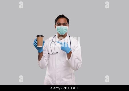 Indian Man Doctor Pointing at Coffee Take Away Cup Wearing Medical Mask and Gloves Isolated. Indian Doctor Holding Coffee To Go Cup. Stock Photo