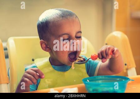 The boy 2 years eats porridge. Children's table. The concept of the child's independence. Cute toddler boy with blue spoon is yogurt. the child smiles Stock Photo