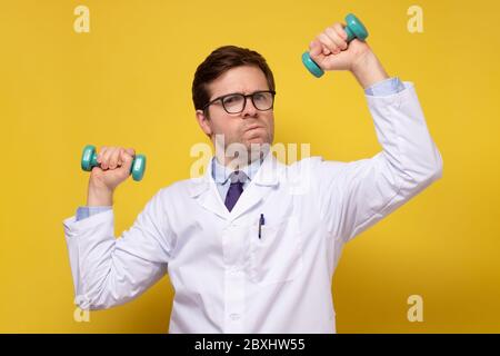 Healthy young male doctor doing exercise using dumbbell. Studio shot on yellow wall. Stock Photo