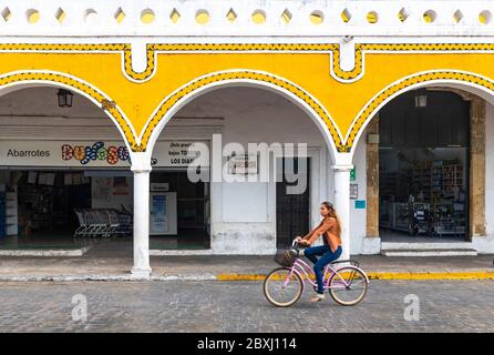 Young mexican woman on a bicycle with blur motion in the yellow city of Izamal with arch architecture by the main square, Yucatan Peninsula, Mexico. Stock Photo