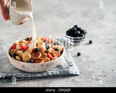 Trendy food - pancake cereal. Milk pouring on heap of mini cereal pancakes in bowl. Tiny cereal pancakes with berries in craft plate over gray cement background. Copy space for text or design. Stock Photo