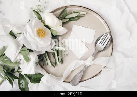 Festive summer wedding scene. Marble table setting with cutlery, olive branches, white peony flowers, stoneware plate and silk ribbon. Blank place car Stock Photo