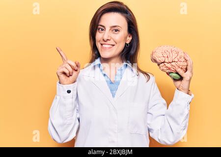 Young beautiful woman wearing doctor coat holding brain smiling happy pointing with hand and finger to the side Stock Photo