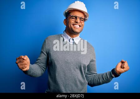 Young handsome african american engineer man with dreadlocks wearing safety helmet very happy and excited doing winner gesture with arms raised, smili Stock Photo
