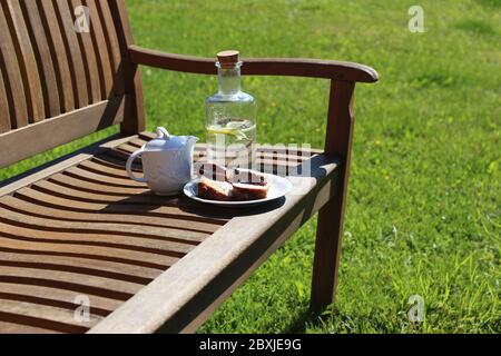 Coffee pitcher, glass bottle of homemade lemonade and cake on wooden bench, blurred green grass background. Outdoor coffee break. Picnic, garden party Stock Photo