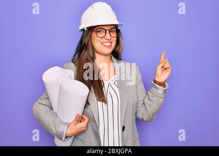 Young hispanic architect woman wearing builder hat and holding blueprints over yellow background very happy pointing with hand and finger to the side Stock Photo