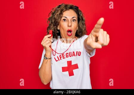 Middle age senior summer lifeguard woman holding whistle over red background Pointing with finger surprised ahead, open mouth amazed expression, somet Stock Photo