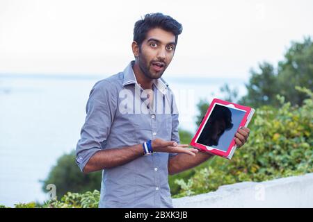 Upset man astonished surprised by what he saw on his tablet, showing its pad screen while standing outside in front of the sea and city park Stock Photo