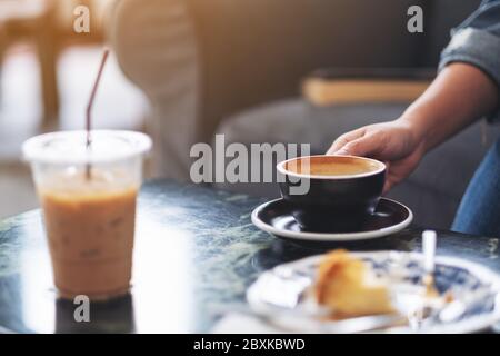 Closeup image of a hand holding a cup of hot coffee with plastic cup and dessert on table in cafe Stock Photo