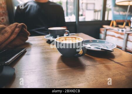 Closeup image of two blue cups of hot latte coffee on wooden table with a man sitting in background Stock Photo