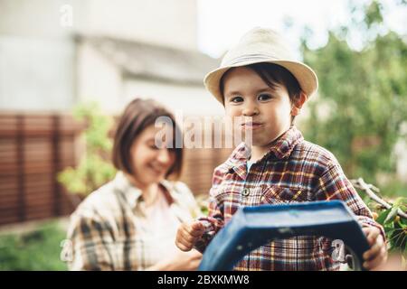 Charming caucasian boy with a hat on head eating cherries from the tree posing with his mother on background smiling Stock Photo