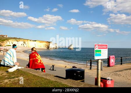 View of Freshwater Bay Isle of Wight two women sat on seafront promenade wall wearing colourful towels chatting paddle board air pump kayak in bay Stock Photo