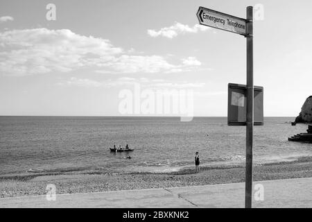 Woman watching kayak carrying three people leaving from the beach shoreline as she stands and watches a swimmer swims by Stock Photo