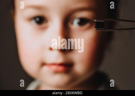 A pair of tweezers holds a black identification microchip against the background of the face. The concept of chipping people after the coronavirus epidemic. Stock Photo