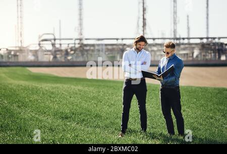 Two young engineers standing outdoors by oil refinery, discussing issues. Stock Photo