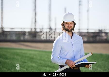Young engineer with hard hat standing outdoors by oil refinery. Stock Photo