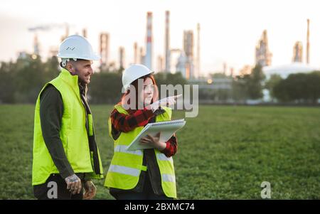 Two young engineers standing outdoors by oil refinery, discussing issues. Stock Photo