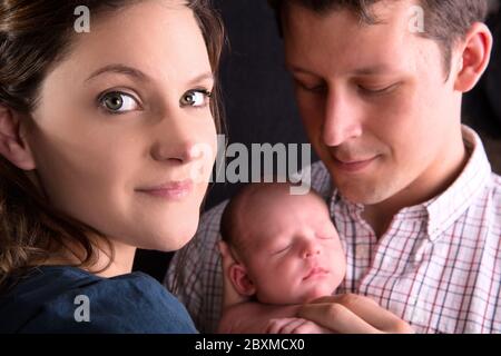 Proud new mother in the foreground smiles to the camera, whilst father holds newborn in the background. Stock Photo