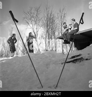 Winter in the 1940s. A young couple is having a cup of coffee while sitting on the roof of their cottage. Their two friends are standing on skis and chatting. The picture is taken in Swedish Lapland near Björkliden. It's Mr and Mrs Iris and Bertil Thorelli having a nice time in the Swedish wilderness.   Sweden 1943 Photo Kristoffersson Ref D115-1 Stock Photo