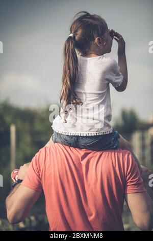 Father holding his daughter on the shouldders and standing on nature summer background. Family or fathers day concept. Back view. Stock Photo