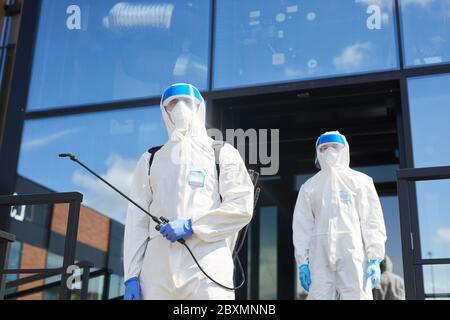 Low angle view at two workers wearing protective suits posing with disinfection gear outdoors while standing against glass building, copy space Stock Photo