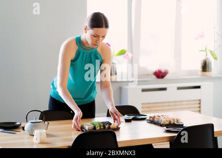 Young woman waiting for guests, friends while arranging the table with various sushi sets in a sunny home with window in the background Stock Photo